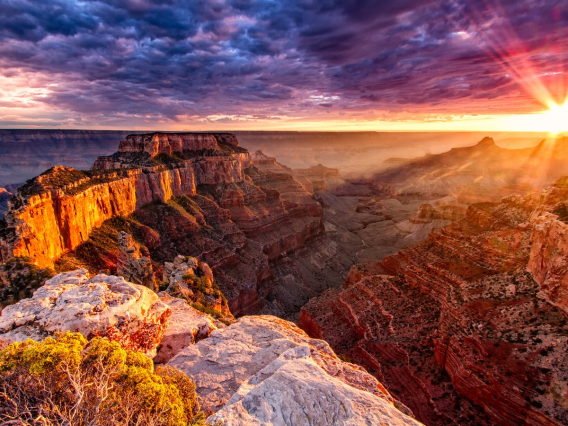 Photograph of the Grand Canyon at sunset with purple clouds overhead and the sun shining through.
