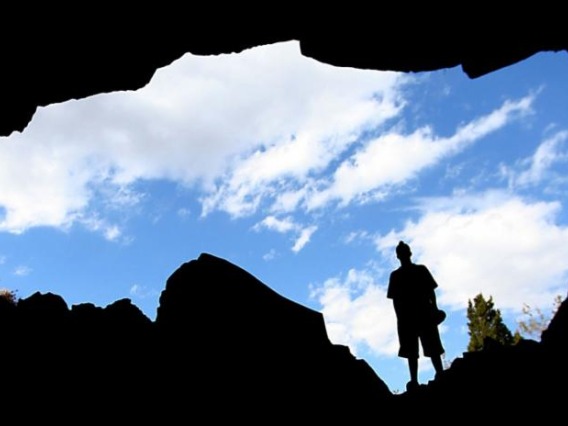 A silhouette of a man standing over an open crevice in the earth with a cloudy blue sky behind him.