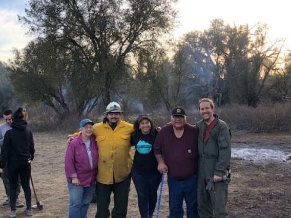 5 people standing next to each other with smoke from a controlled fire in the background.