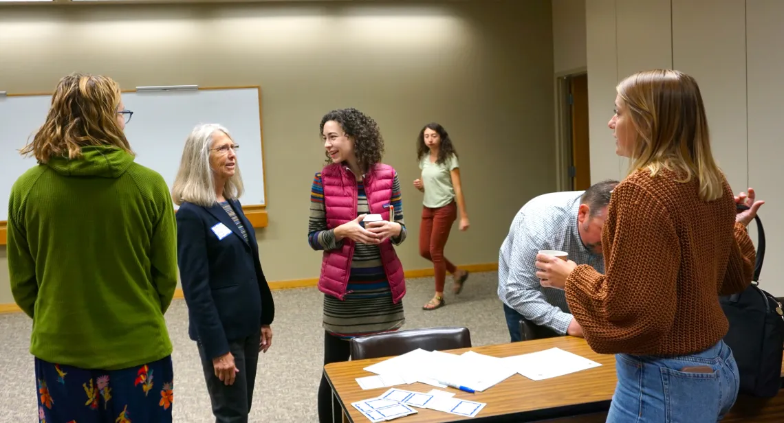 4 People stand around a table with documents on it, one person walking in the background
