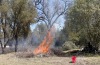 a controlled fire in a wooded area during daylight. The fire is burning a pile of dead branches and leaves, with flames reaching high into the air. In the foreground, there is a red bucket and a tool, possibly a rake, lying on the ground. Trees surrounding the fire appear healthy and unaffected by the flames. Smoke is visible, rising from the fire and blending with the clear sky above.